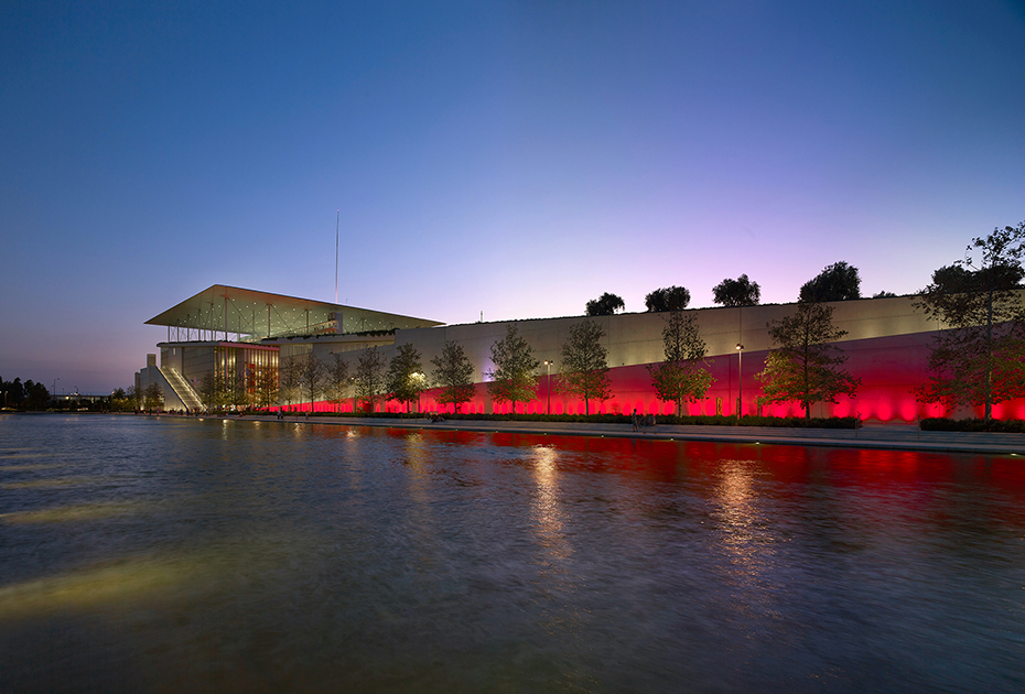 Photo showing a pond at the end of which there is an illuminated wall and a large glass building at the edge