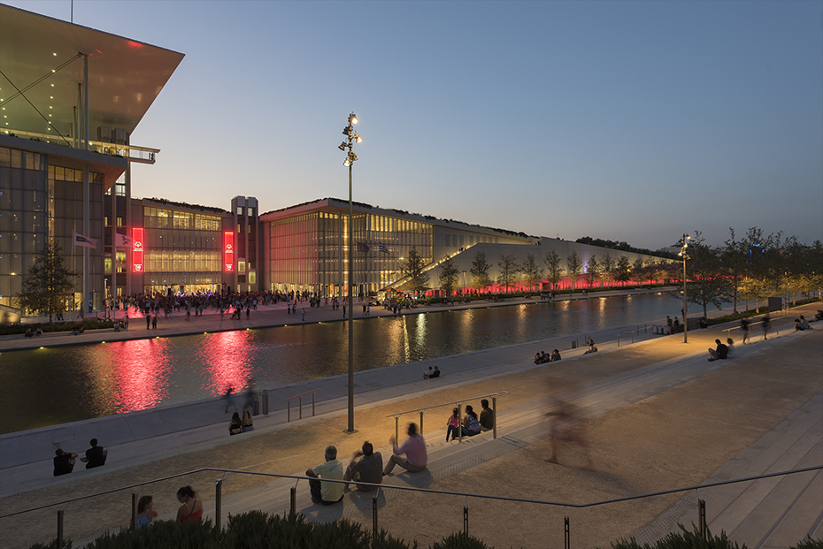 Photo showing people sitting on steps looking at a lake and a large glass building in front of them