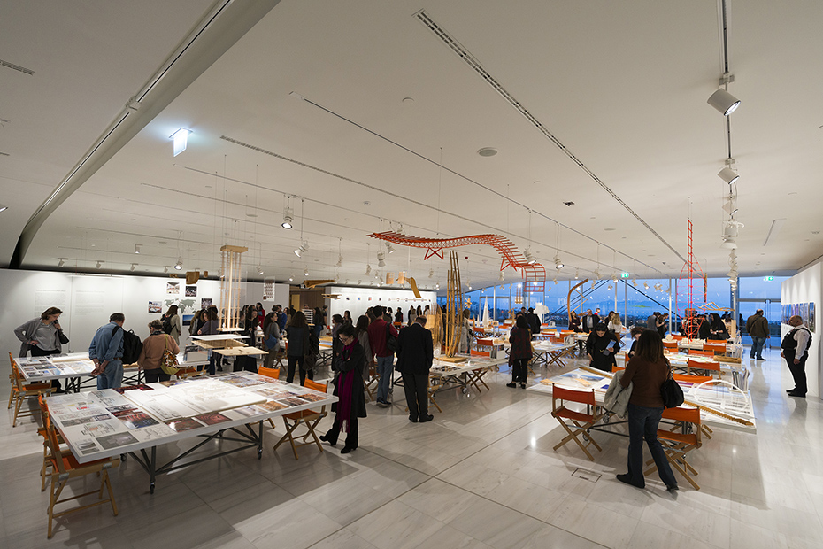 Distant photograph showing people standing and looking at tables on which there are drawings, memos, pictures and models of buildings