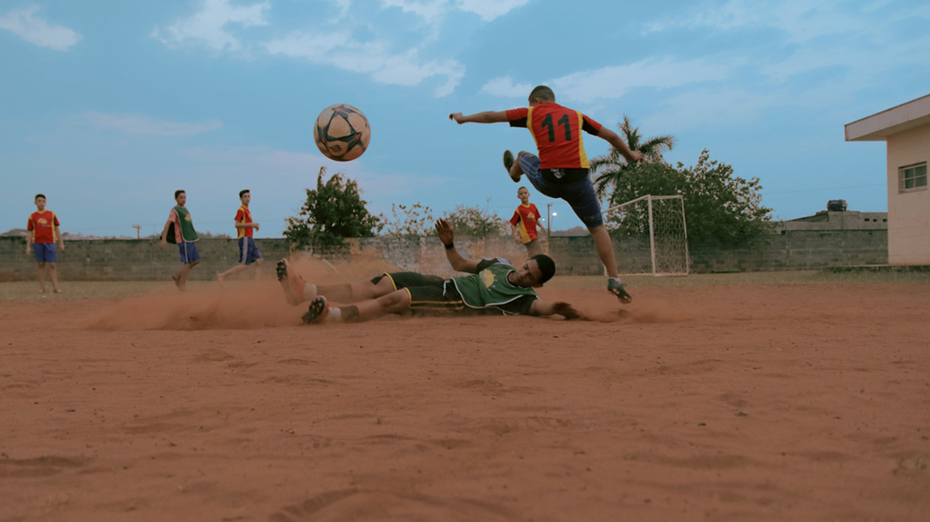 A group of children are playing football and one boy is lying on the floor while his opponent jumps on top of him