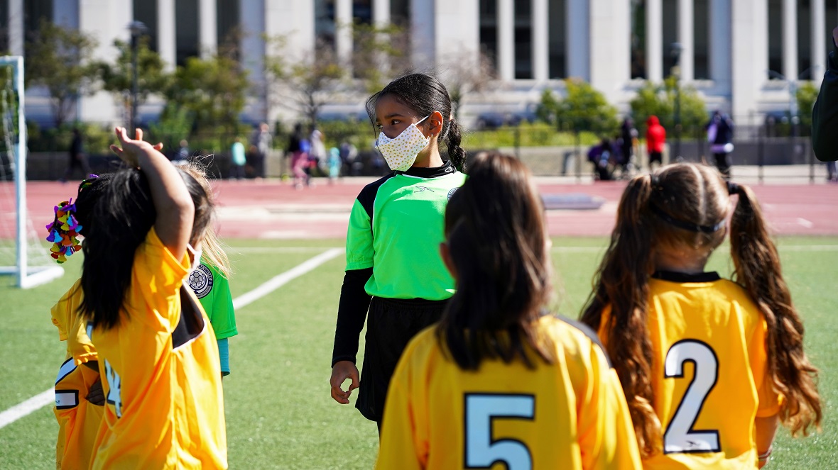 Little girls wearing yellow football uniforms are gathered together and listening to the girl who has the role of the keeper wearing a green football uniform