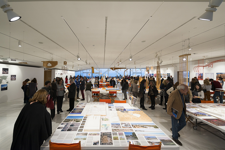 Distant photograph showing people standing and looking at tables on which there are drawings, memos, pictures and models of buildings