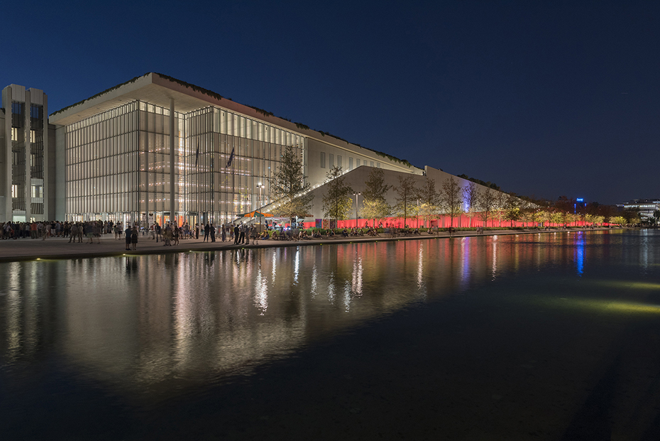 Photo taken at night showing a crowd of people walking on the sidewalk between a lake and a large glass building