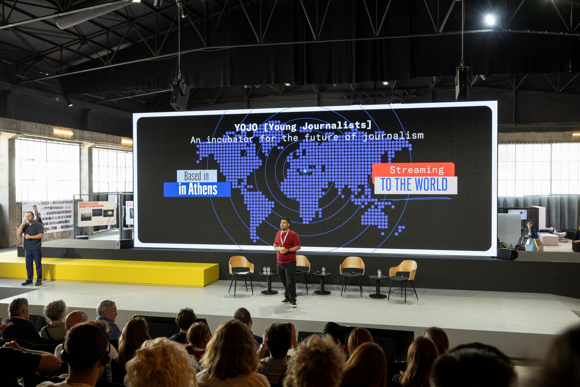 A speaker presents on stage with a large screen reading