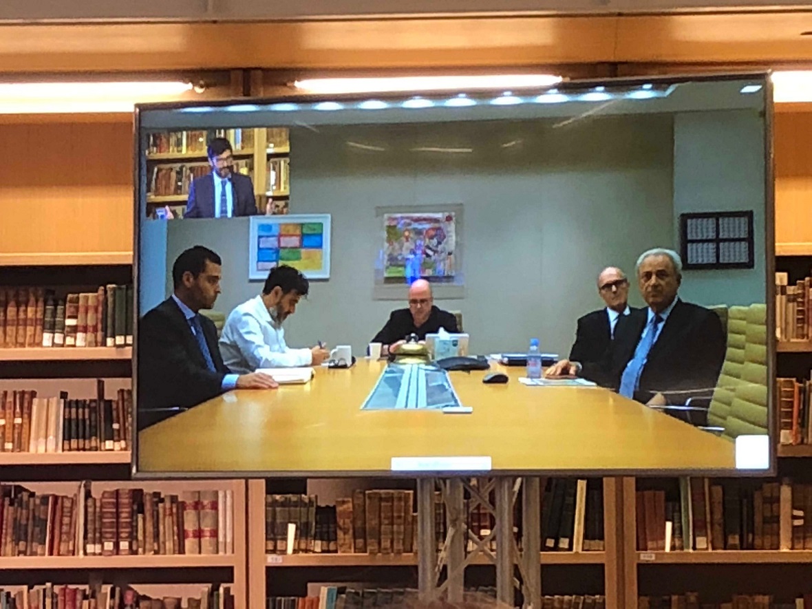 A photo showing the screen on which a live connection is displayed. On one side you can see five men sitting around a conference table and on the other side you can see a man talking