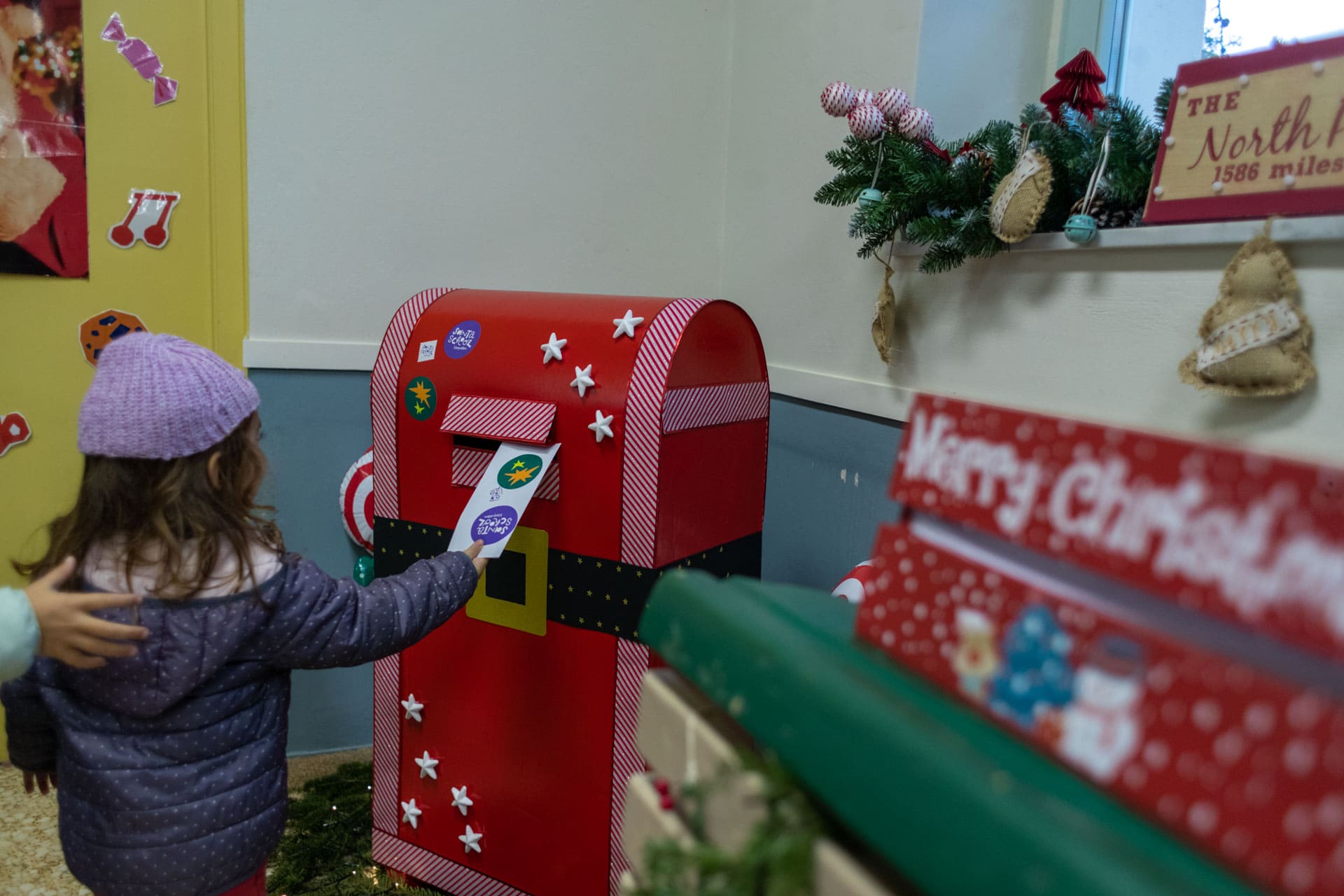 A child sending a Christmas letter in a red post box, inside a room.