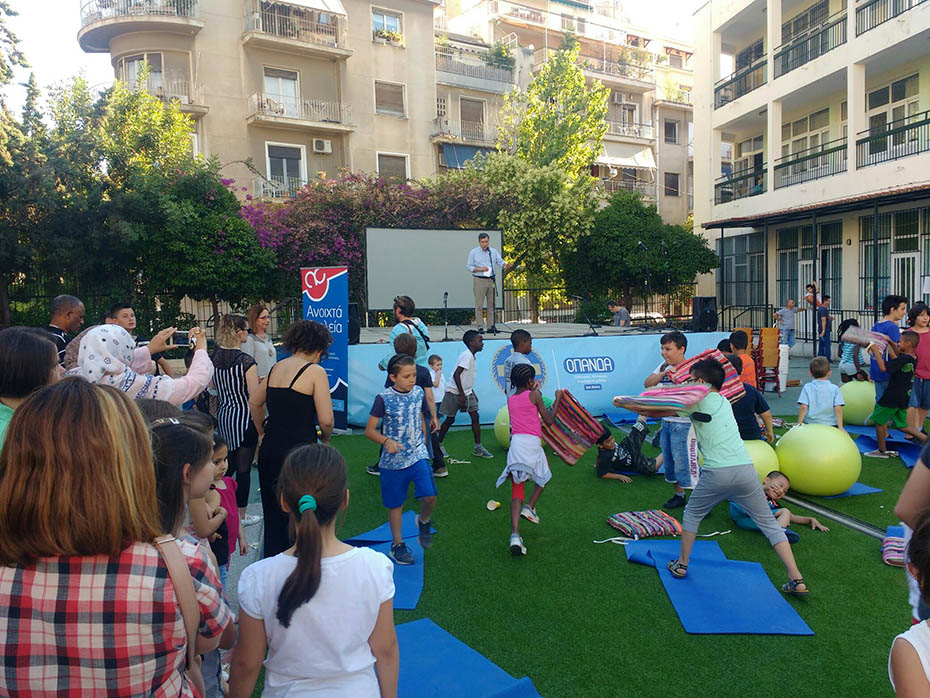 In the courtyard of a school there is a stage and grass and children play while people watch them