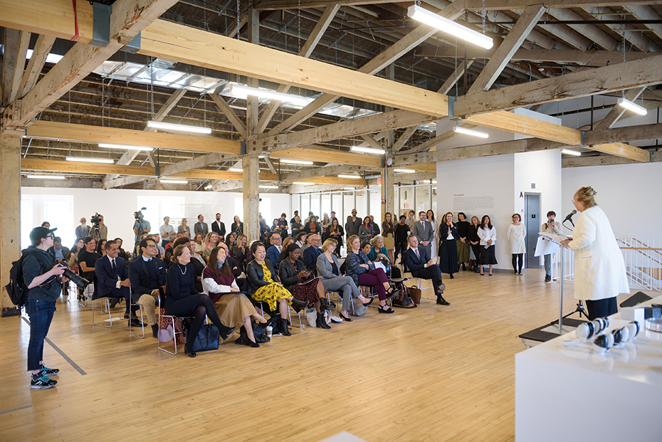 A woman gives a speech to a seated crowd inside an art center