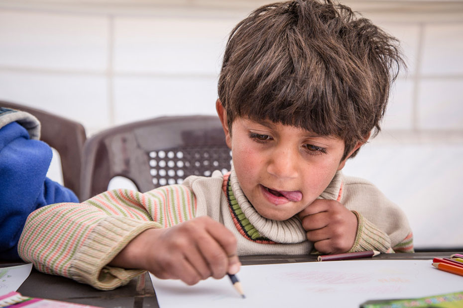 A little boy sits at the table and draws with his tongue out