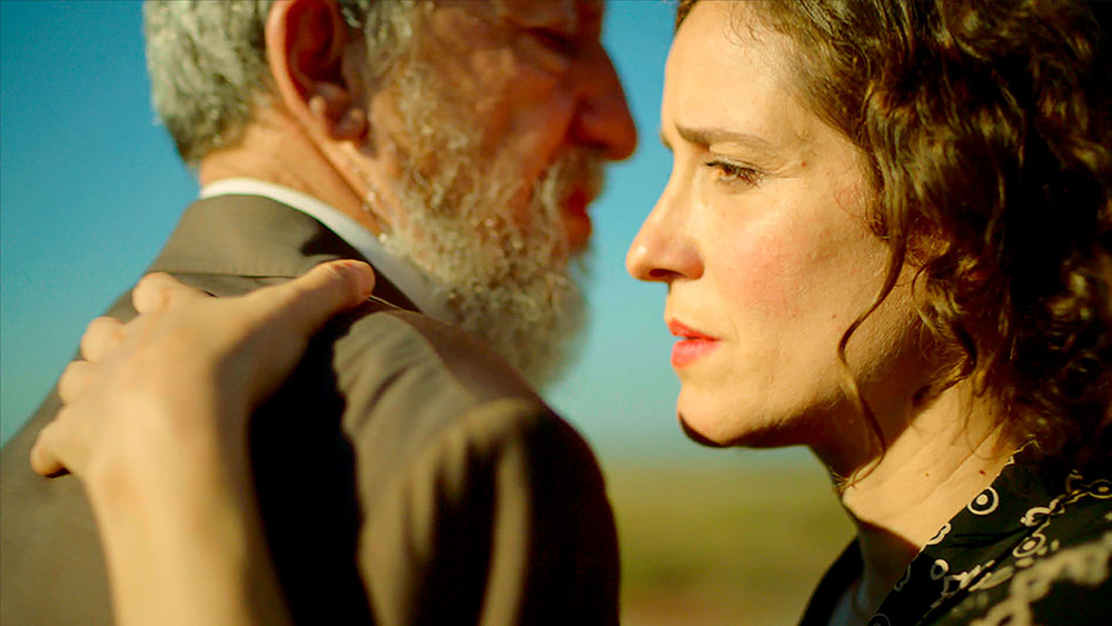 A woman and a man stand facing each other and the woman touches him on the shoulder