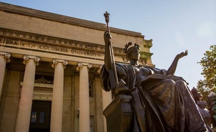 A photograph of a bronze statue of a woman reading a book with her arms spread outside of the Library of Columbia University