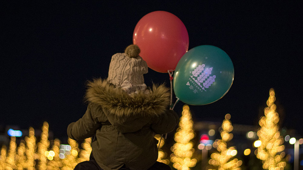 A child sitting on their parent's shoulders looks onto the decorated trees, while holding two balloons