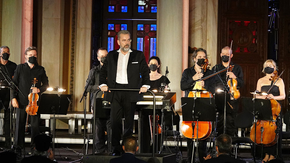 On a stage an orchestra stands upright with its members dressed in black and in front the conductor looks at the audience