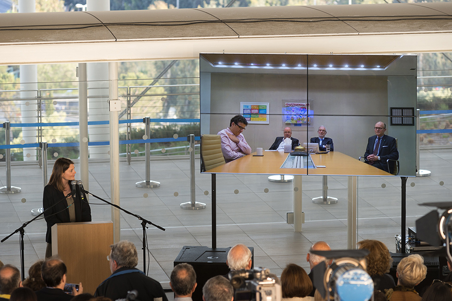 A photo showing a woman on the stage addressing the four men who are participating via an internet connection and the audience watching them