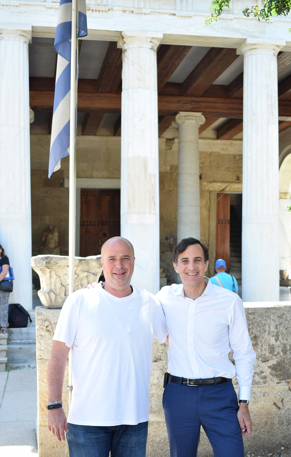 Ο Ανδρέας Δρακόπουλος και ένας άλλος άντρας χαμογελούν και κουνιούνται και βγάζουν μια φωτογραφία έξω από ένα αρχαιολογικό μουσείο με την ελληνική σημαία στο φόντο
