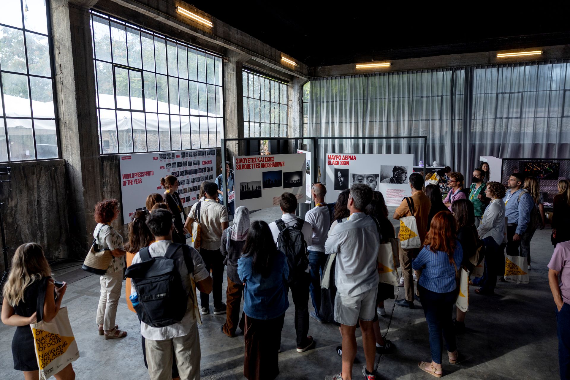 A group of people views photo exhibits in a room with large windows.