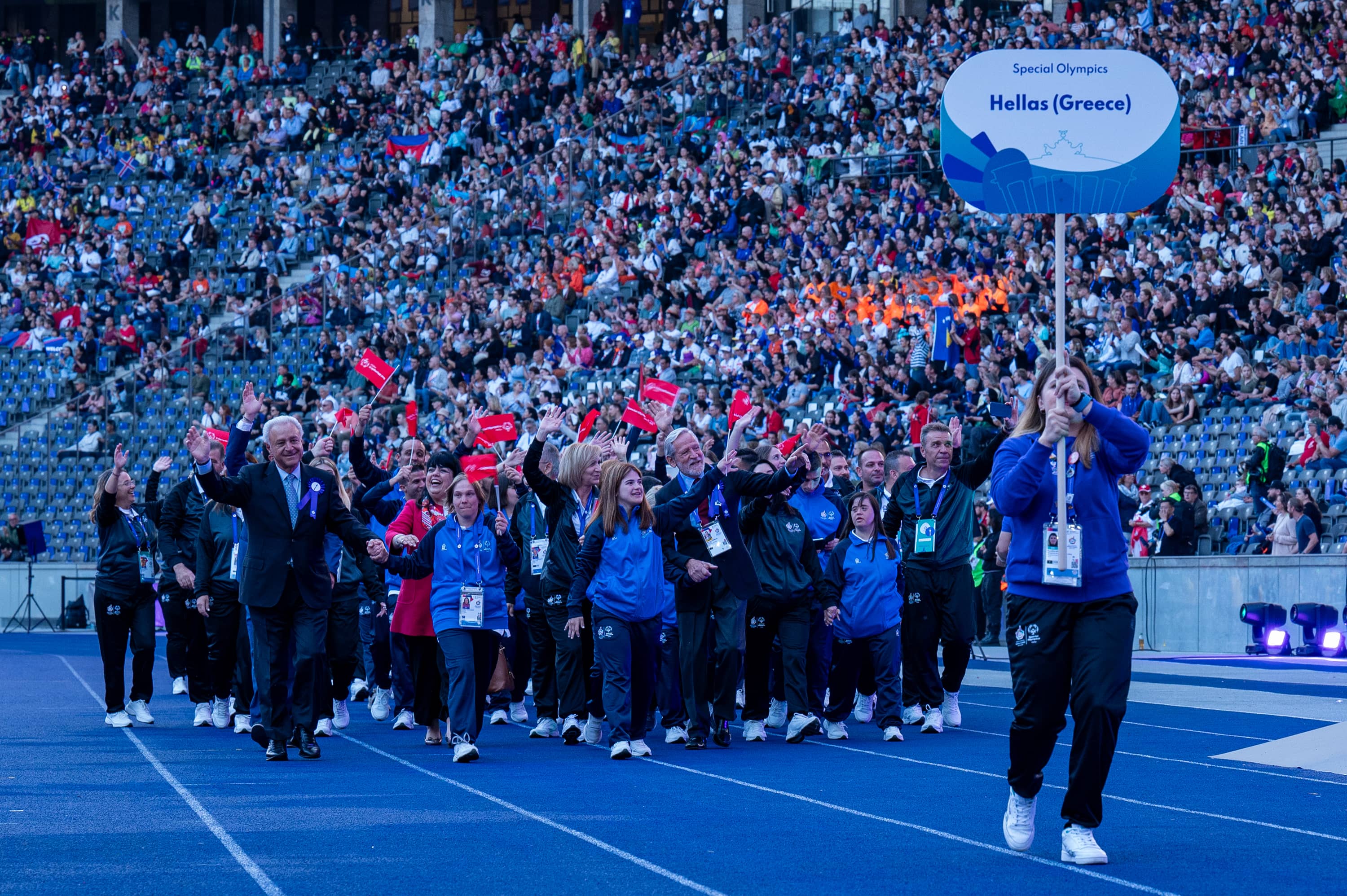A woman holds the Hellas sign in front of Special Olympic athletes who are waving and walking together on the track