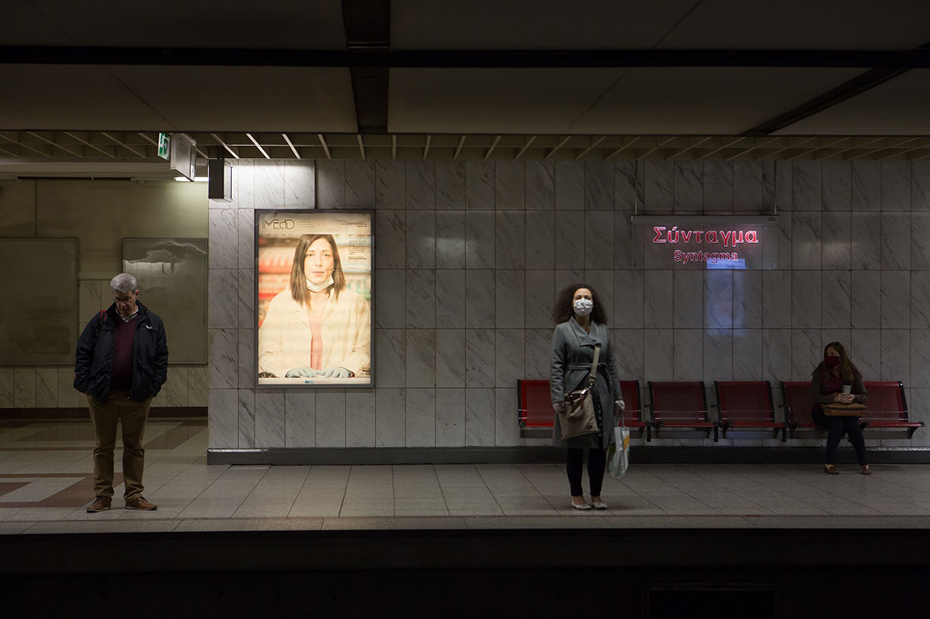At Syntagma metro station there is a man and a woman standing up, a woman sitting down and a screen showing a vulture and the iMEdD logo