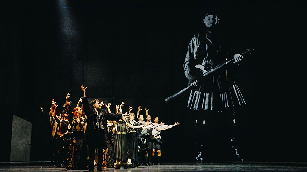 On a stage a group of actors dressed in Greek war uniforms stand and show with their hands a photograph of a man from the war standing with his gun