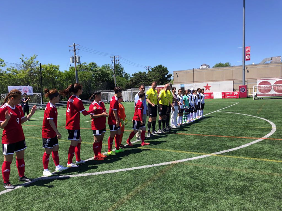 On a football field, two women's football teams are standing on the center line and between them two referees
