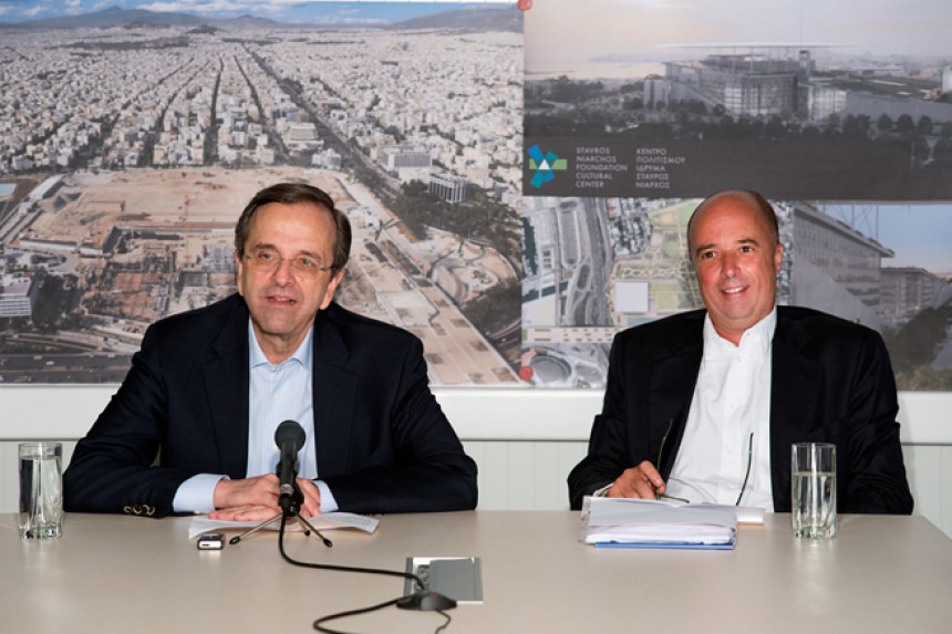 Antonis Samaras, the Prime Minister of the Hellenic Republic, and Andreas Dracopoulos, the co-president of the Stavros Niarchos Foundation, sit a table in front of a picture of the construction site of the Stavros Niarchos Foundation Cultural Center