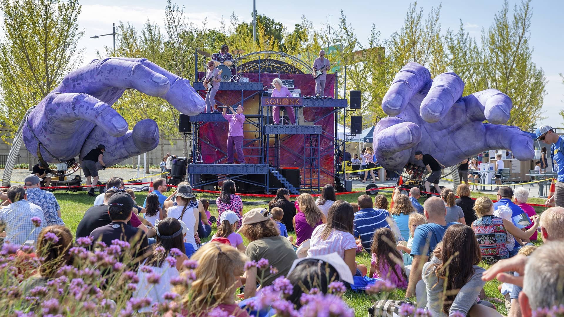 A crowd of people sit on the grass and watch a concert where the stage is decorated with two big purple hands