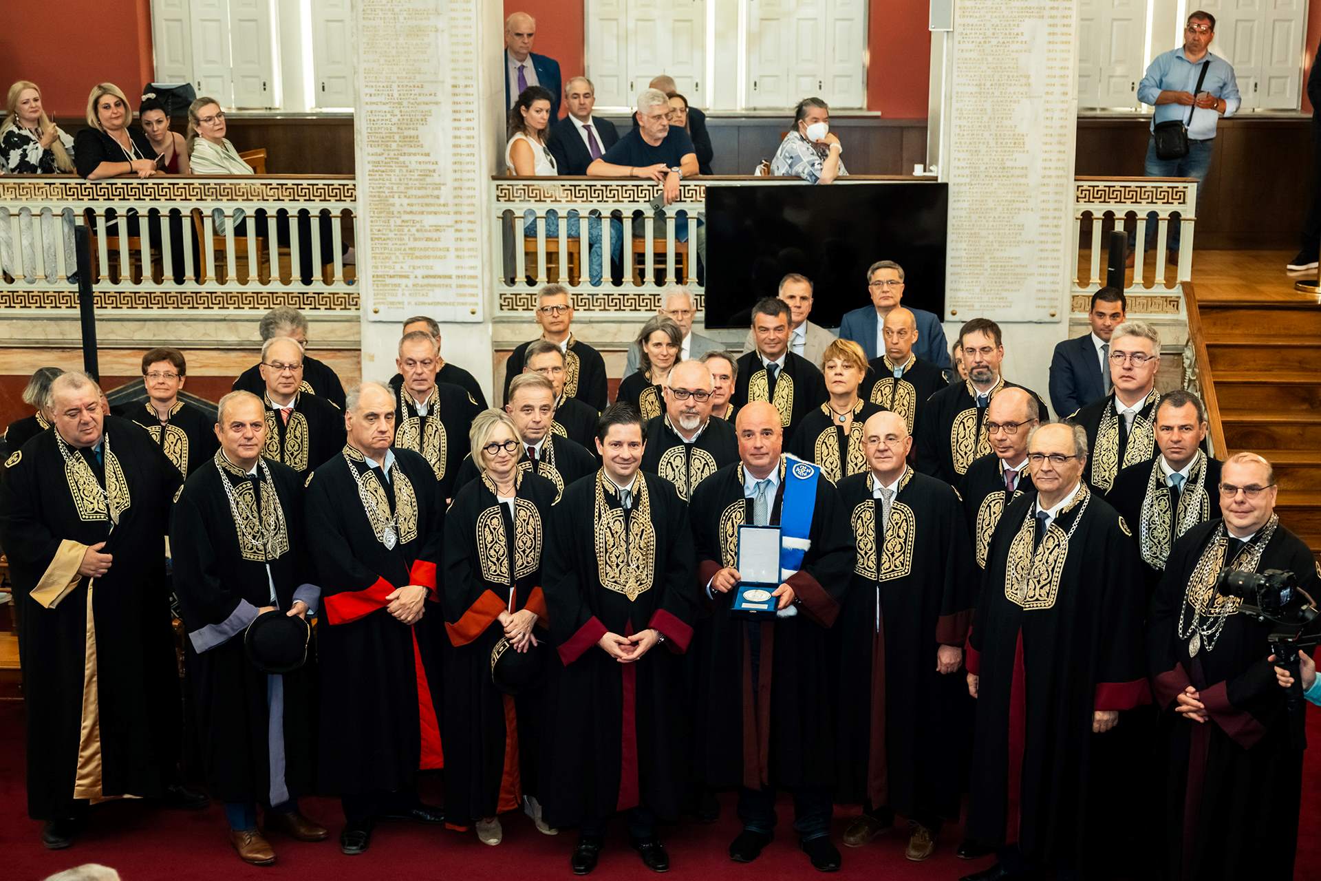 The honored SNF Co-President, Andreas Dracopoulos, holding an honorary plaque, standing among members of the Senate and the academic community of the University of Athens. All of them are dressed in gowns.