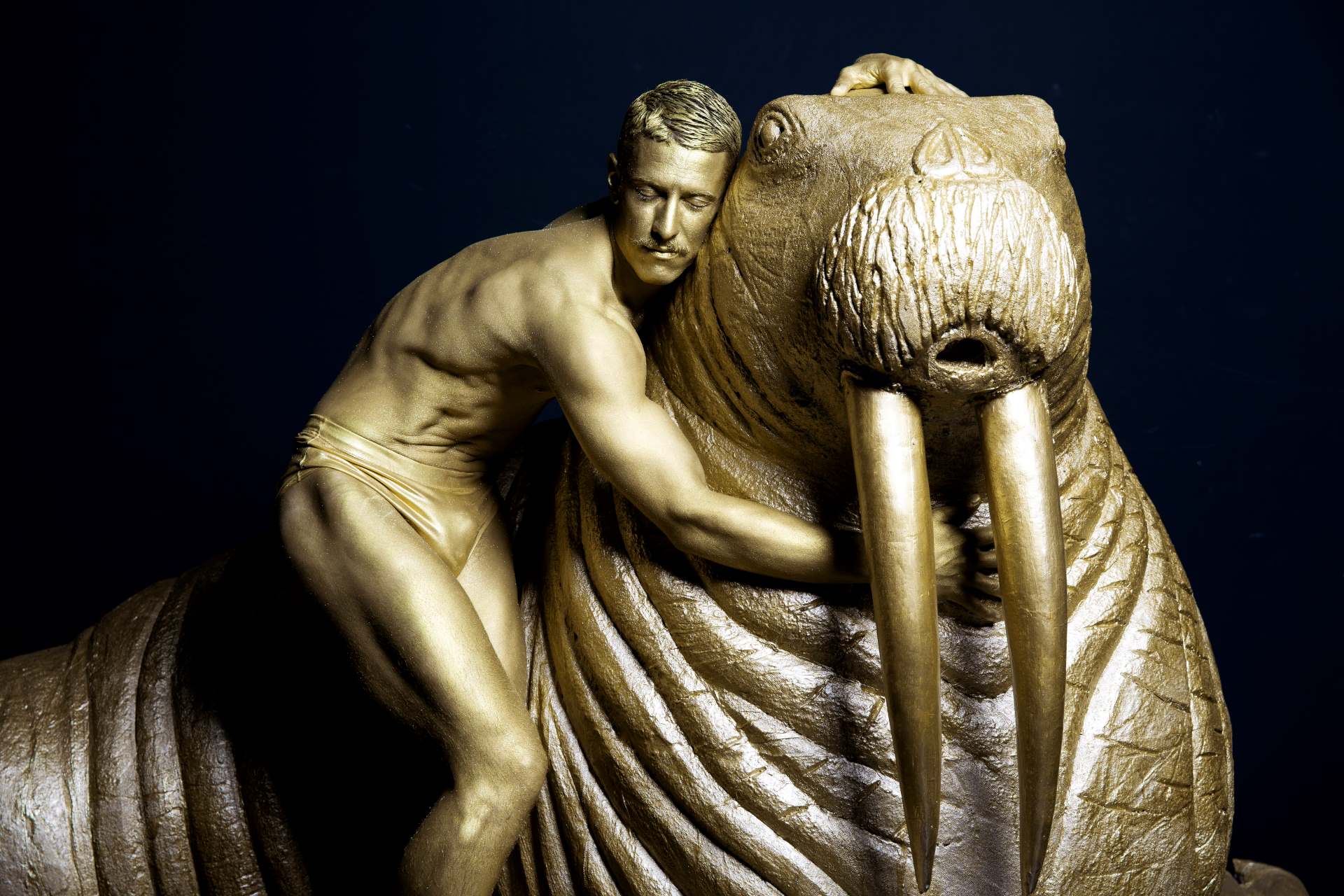 Man hugs walrus while both of them are painted gold.