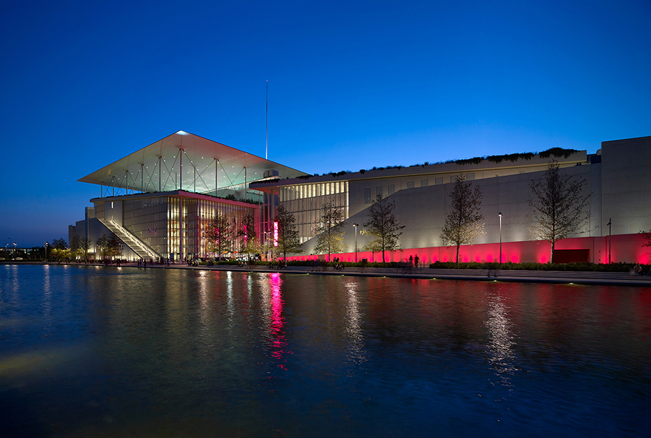 Photo showing a pond at the end of which there is an illuminated wall and a large glass building at the edge