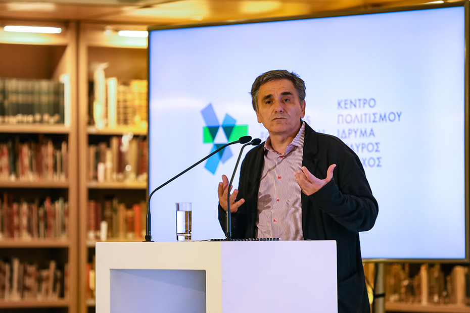A man stands on the podium and speaks while behind him a photograph is projected which reads ΚΕΝΤΡΟ ΠΟΛΙΤΙΣΜΟΥ ΙΔΡΥΜΑ ΣΤΑΥΡΟΣ ΝΙΑΡΧΟΣ