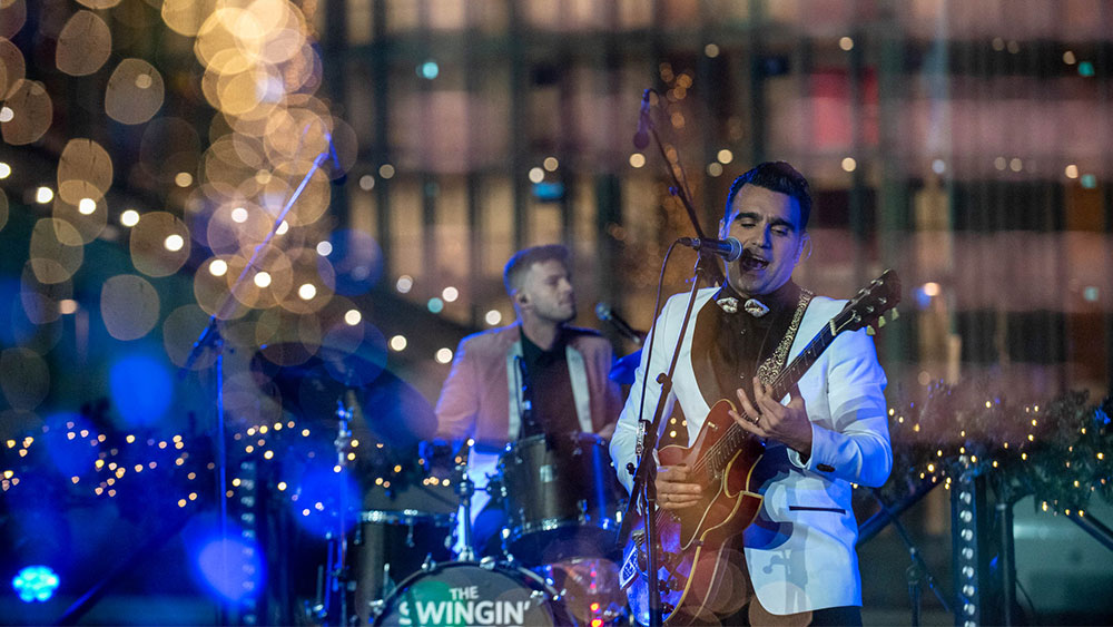 A man sings and plays the guitar, while another man plays the drums in the back