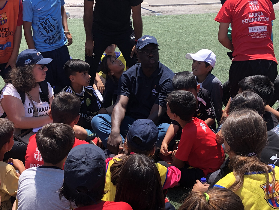 A man and a group of young children sit squatting on a football field and discuss