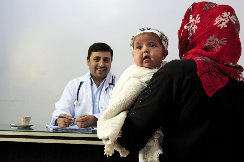 A doctor sits at his desk and smiles at a mother holding her baby