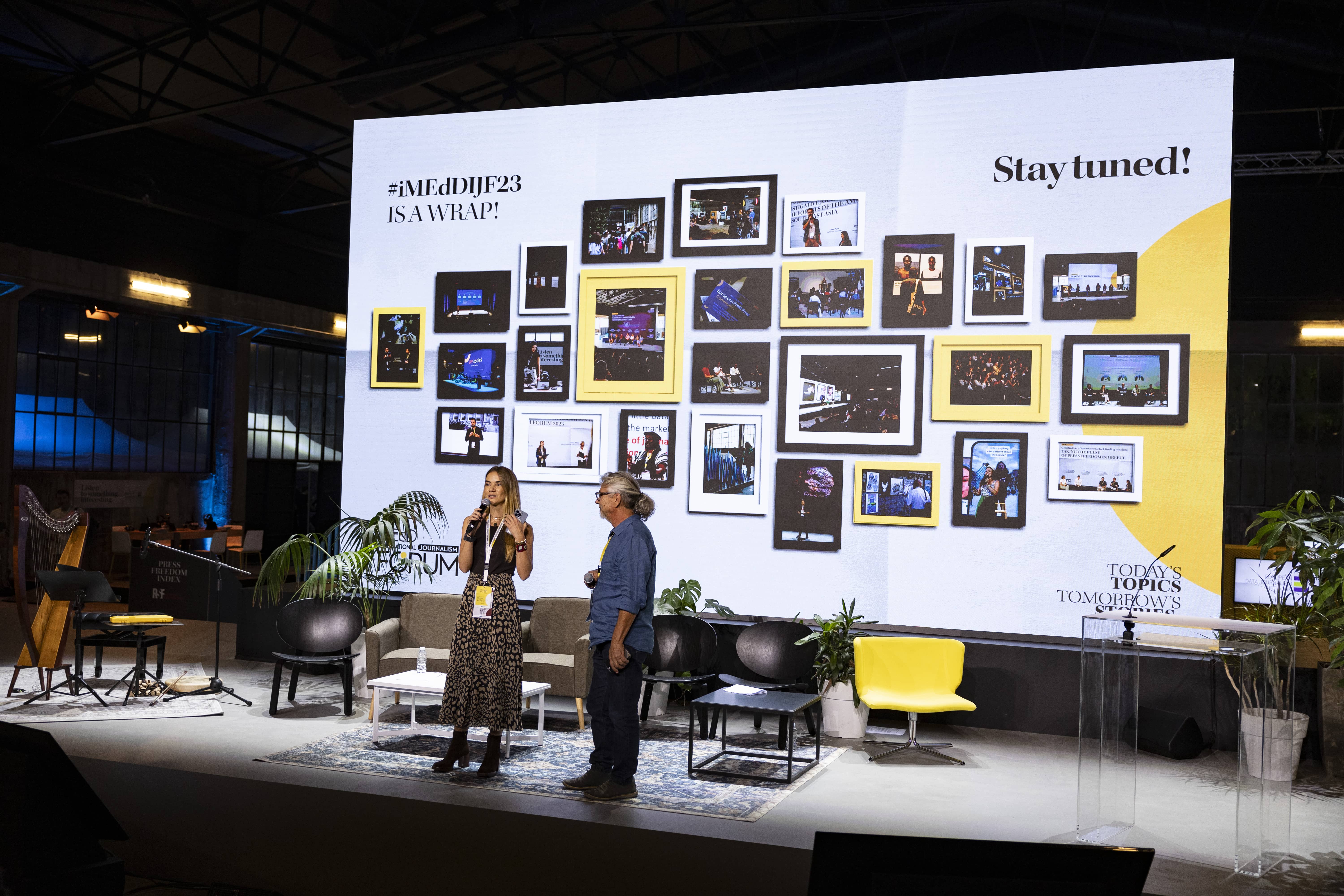 Anna - Kynthia Boussdoukou and Stratis Trilikis, founders of iMedD, stand on a stage where there are chairs and a large illuminated screen which shows many frames with photos and says iMedDJF2023 its a wrap stay tuned and talk to the audience watching them 