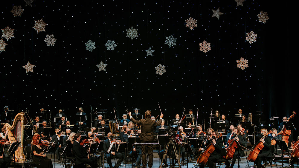 A man conducts an orchestra in front of snowflake decorations