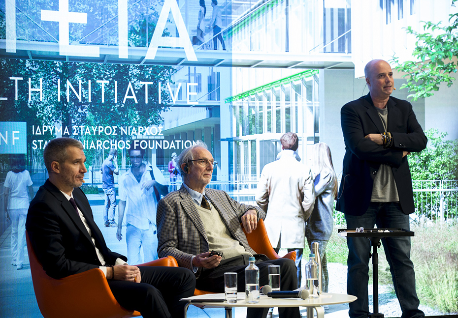 Andreas Dracopoulos, President of the Stavros Niarchos Foundation, is standing on stage and next to him are two other men sitting