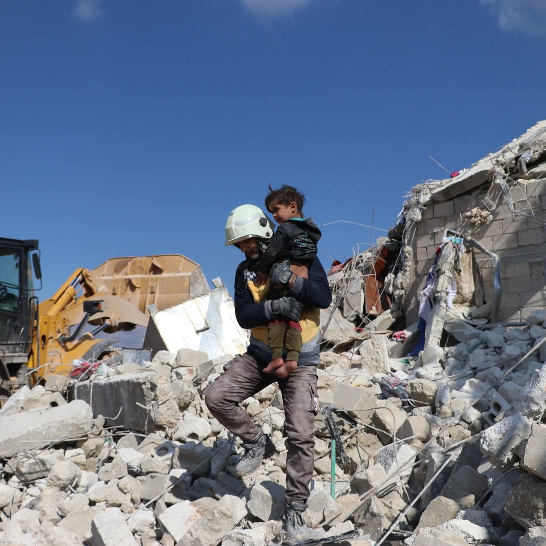 An adult in a white helmet carries a child down from a pile of rubble.