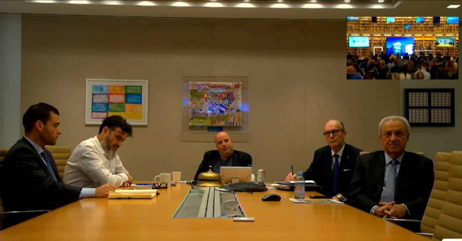 Photo via a live connection showing five men sitting around a large conference trapeze and communicating with an audience sitting in chairs