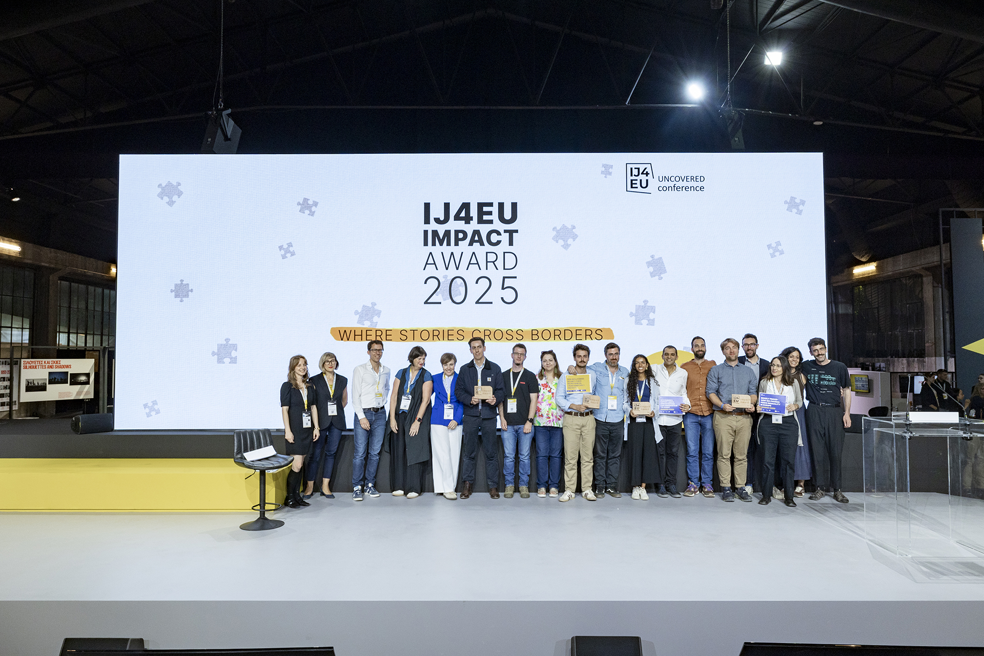 A group of award recipients stands on stage holding certificates in front of a screen reading