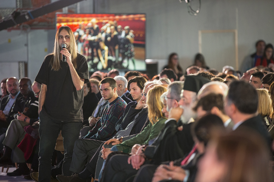 A young man from the audience stands up and raises a concern at the microphone as the rest of the audience looks on