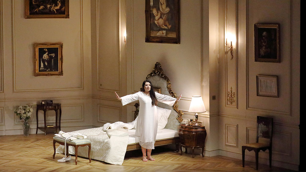 A woman standing upright with her arms open wearing a white nightgown in front of a bed in a large and emblematic room