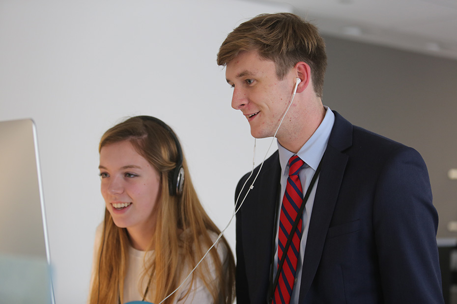 A young man and a young woman standing next to a computer screen and wearing headphones