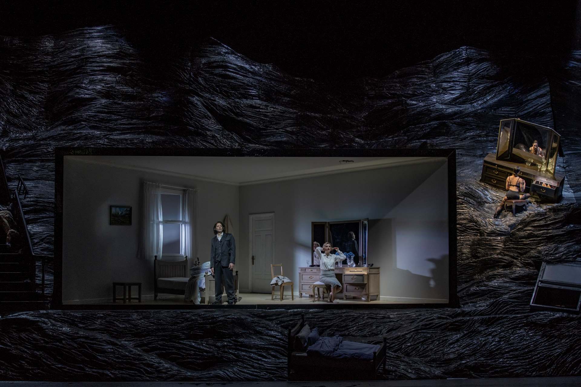 In a room, a man stands in front of the bed while a woman stands in front of a mirror.
