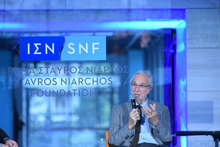 A man sits on stage and speaks holding a microphone. Behind him is a blue background that reads ΙΔΡΥΜΑ ΣΤΑΥΡΟΣ ΝΙΑΡΧΟΣ STAVROS NIARCHOS FOUNDATION
